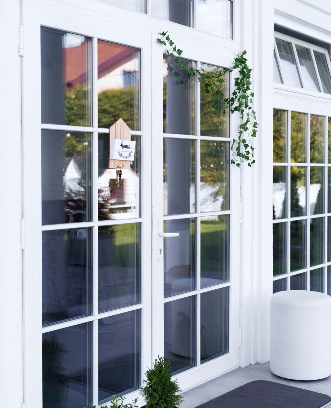 Partial view of double glass doors and a tall window. The doors and window are each framed in white wood with cascading ivy. A small decorative plaque hanging from the door reads Home.