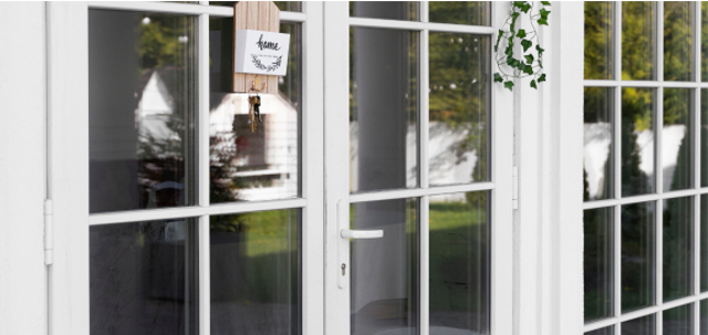 Partial view of double glass doors and a tall window. The doors and window are each framed in white wood with cascading ivy. A small decorative plaque hanging from the door reads Home.